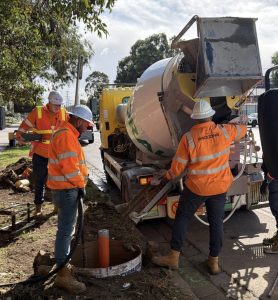 Construction crew in orange safety vests and hard hats pour concrete into an open manhole beside a cement mixer truck on a sidewalk.
