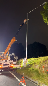 Nighttime roadside maintenance: an orange crane extends to a tall streetlight pole, with two workers in safety gear near the base and traffic cones nearby, preparing or replacing the lamp.