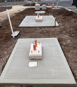 Construction site with three concrete utility pads, orange‑capped pipes, and surrounding dirt in a street-side trench area.