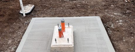 Construction site with three concrete utility pads, orange‑capped pipes, and surrounding dirt in a street-side trench area.