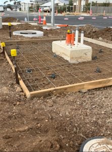 Construction site with a reinforced steel rebar grid in wooden formwork, utility pipes protruding from a concrete block at center, on a dirt lot. nearby street in background