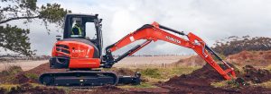 Orange Kubota mini excavator digging a trench in a rural field, operator in high-visibility vest.